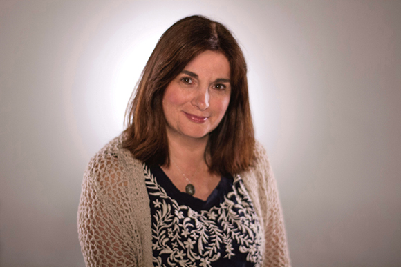 A woman with brown hair wearing a patterned navy top and a light knit cardigan smiles against a grey background.
