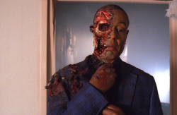 A man in a blue suit with bloody, decaying flesh on half of his face and his hand.