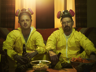 Two men wearing yellow hazmat suits and goggles sit on a couch with beer and snacks.