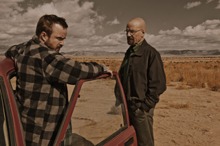 A man in a plaid shirt leans on a car door while facing a bald man in a jacket in a desert.