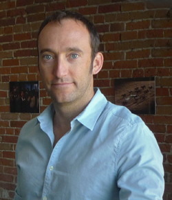 A man with short brown hair wearing a light blue button-down shirt stands before a brick wall.