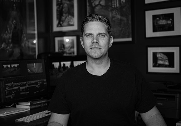 Black and white portrait of a man in a black t-shirt sitting in front of framed pictures and computer monitors.