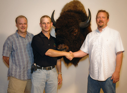 Three men stand in a row, with the middle man shaking hands with the man on the right in front of a bison mount.