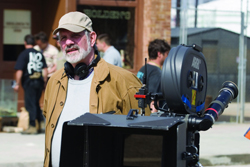 A man with a beard and cap stands behind a large motion picture camera on an outdoor film set.