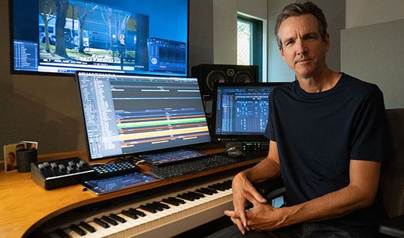 A man sits at a desk with three computer monitors, a MIDI keyboard, and audio mixing equipment.