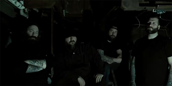 Four bearded men in dark clothing pose in a dimly lit, shadowy indoor environment.