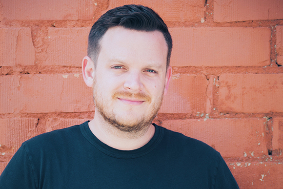 A man with short dark hair and a beard wearing a black t-shirt, smiling in front of a red brick wall.