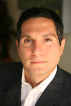 A man with short dark hair wearing a white shirt and a dark pinstripe suit jacket.