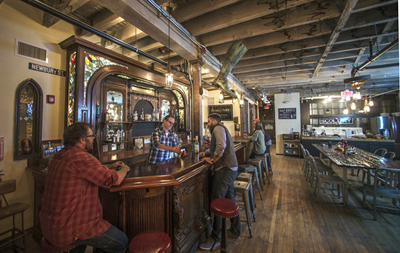 People sit at a long wooden bar inside a rustic pub with exposed ceiling beams and wooden floors.
