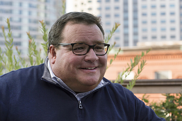 A smiling man with dark hair and glasses wearing a navy blue quarter-zip pullover outdoors.