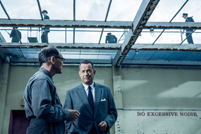 A man in a suit talks to a man in a grey shirt in a courtyard with guards on a walkway above.
