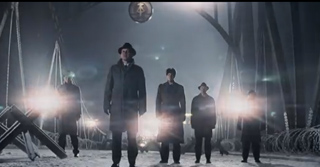 Five men in dark coats stand in a line within a dark, foggy industrial setting with bright lights.