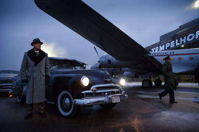 A man in a grey coat and hat stands by a black vintage car next to a large propeller plane at night.