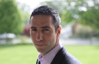 A man with short dark hair wearing a light pink shirt and dark blazer outdoors.