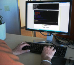 Person's hands typing on a black keyboard in front of a computer monitor showing lines of code.