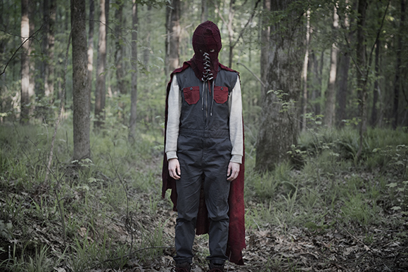 A person wearing a dark red hooded mask and grey overalls stands in a dense, wooded area.