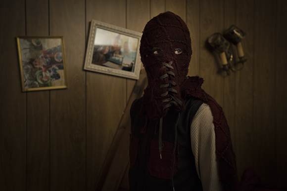 A person in a dark red, textured mask with eye holes and laces stands before a wood-paneled wall.