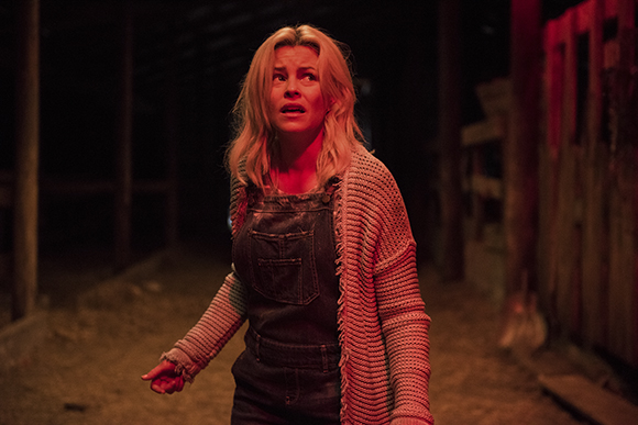 A blonde woman in denim overalls and a grey cardigan looks ahead with a fearful expression in a dark room.