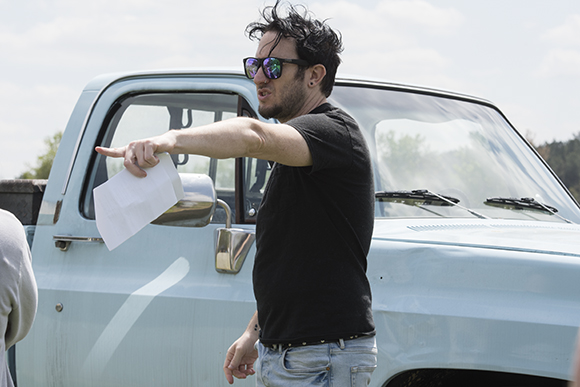 A man in a black t-shirt and sunglasses points while holding a white paper next to a light blue pickup truck.