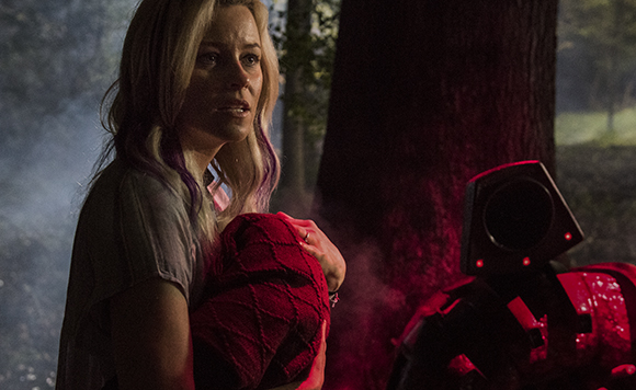 A blonde woman with a concerned expression holds a red blanket in a dark, smoky forest setting.