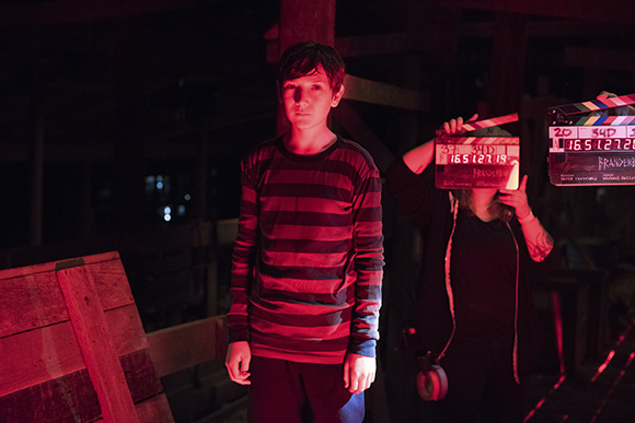 A boy in a striped shirt stands in a dark, red-lit setting next to a person holding a film clapperboard.