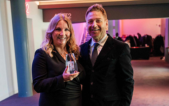 A smiling man in a suit and a woman in a black blazer hold a glass award together.