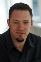 A man with short dark hair and a small goatee wearing a black collared shirt.