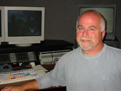 A smiling man with gray hair and a goatee sitting in front of computer monitors and an audio mixing console.