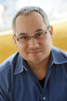 A man with short hair and glasses wearing a blue button-down shirt, smiling at the camera.