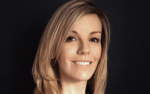 A smiling woman with blonde hair looks toward the camera against a dark background.