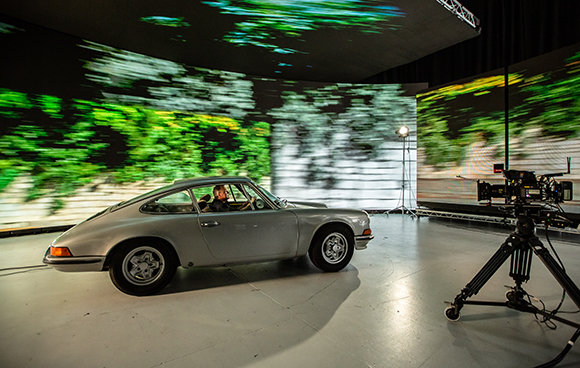 A silver vintage sports car sits in a studio with large curved LED screens displaying a blurred forest landscape.