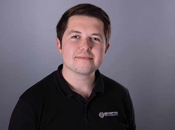 A man with short brown hair wearing a black polo shirt against a gray background.