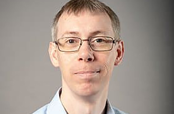 A man with short brown hair and glasses looking directly at the camera against a gray background.