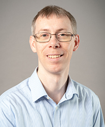 A man with short brown hair and glasses wearing a light blue button-down shirt.