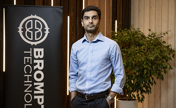 A man in a light blue shirt stands next to a black Brompton Technology banner and a potted plant.