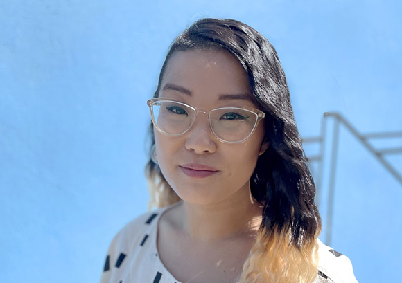 A woman with dark, wavy hair and clear-framed glasses looks forward against a clear blue sky.
