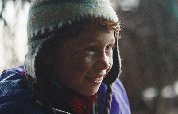 A smiling child with freckles wearing a patterned knit earflap hat and a purple jacket.
