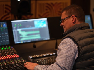 A man wearing glasses and a quilted vest operates a large audio mixing console in a dark studio.