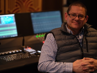 A man with glasses and a grey quilted vest sits before computer monitors and a sound mixing board.