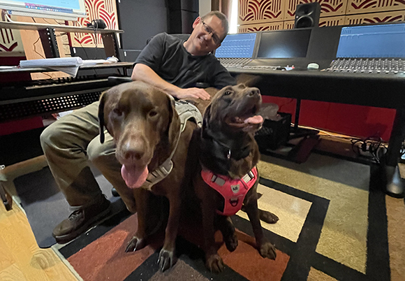 A man sits behind two chocolate Labrador Retrievers in a music recording studio.