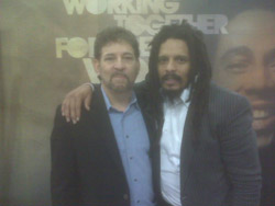 Two men stand together with arms around each other in front of a backdrop with text and a partial image of a face.