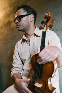A man in a tan shirt and glasses holds a violin vertically against a textured wall.