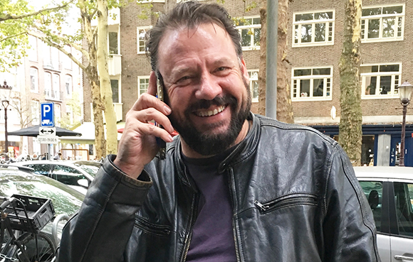 A smiling man with a beard wearing a black leather jacket talks on a cell phone outdoors.