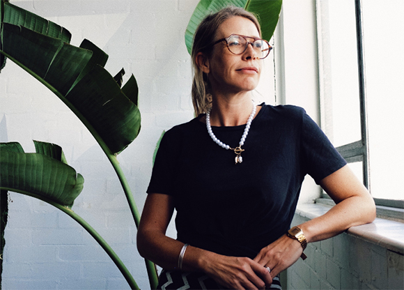 A woman with glasses and a pearl necklace stands by a window next to a large tropical plant.