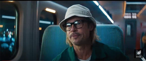 A man with glasses and a white bucket hat sits in a blue striped train seat.