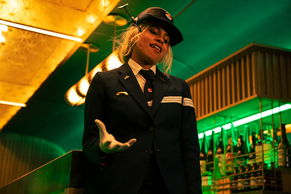 A person in a dark uniform and hat gestures with an open hand in a room with green lighting.