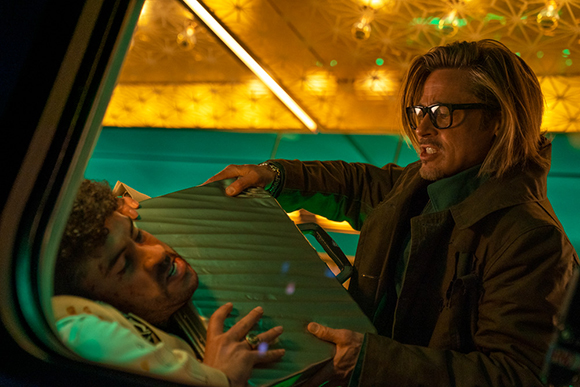 A man with long hair and glasses holds a metal briefcase against a man's face in a dimly lit vehicle.