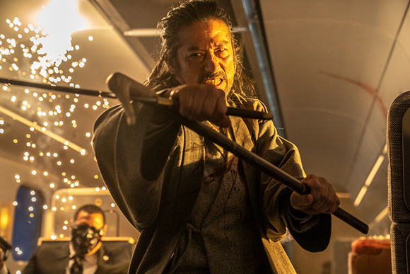 A man in dark robes grips a long staff as bright sparks fly behind him in a dimly lit setting.