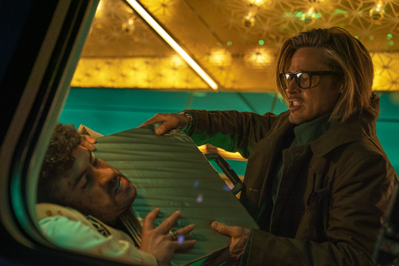 A man with blonde hair and glasses holds a large metal sheet over a man lying down.