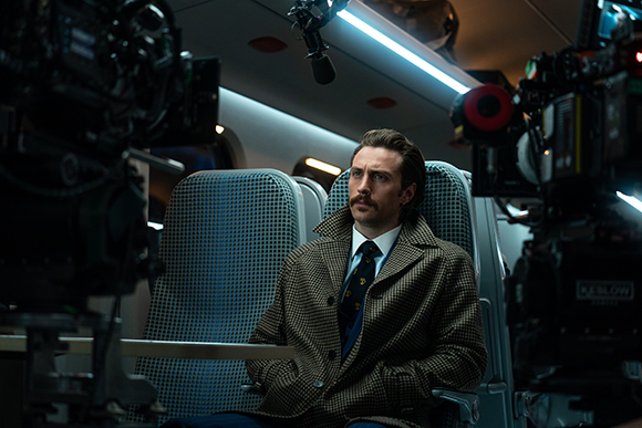 A man with a mustache wearing a brown patterned coat sits in an airplane seat surrounded by film cameras.